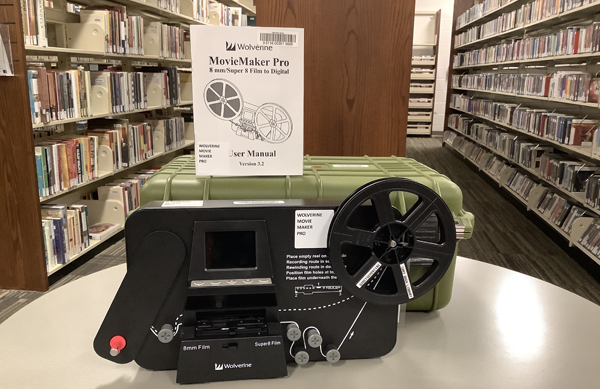 Photo of the Wolverine MovieMaker Pro device in front of it's green case on top of a table with book shelves in the background. The instruction booklet is standing atop the case.