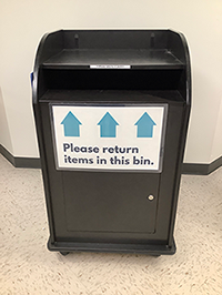 Photo of our black return bin with a sign that reads "Please return items in this bin."