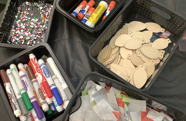 Overhead photo of five small black bins containing art supplies: gems, glue sticks, wooden ornaments that look like light bulbs, tissue paper, and markers.