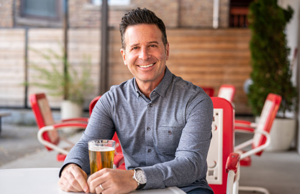 Steve Dolinsky sits at a table with a glass of beer, facing towards the camera and smiling.